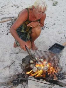 Woman cooking clams over fire on a beach.