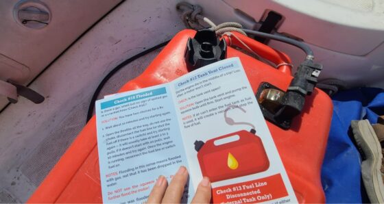 The Boat Galley Outboard Troubleshooting for Beginners pamphlet open on top of a red plastic outboard fuel tank in a dinghy, showing two pages with Check / Solution / Notes sections for "Flooded" and "Tank Vent Closed" problems.