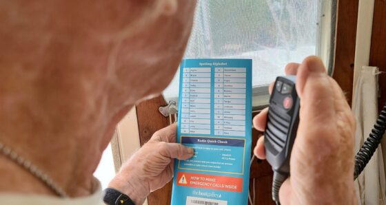 A man's hand holding the Boat Galley Handy VHF Reference card open to the Spelling Alphabet and Radio Quick Check pages, with a black VHF microphone in his other hand below the card.