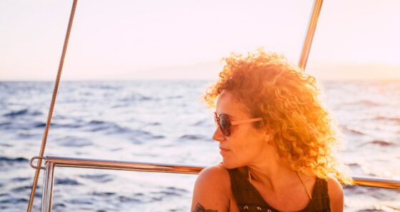 Woman aboard a sailboat at sunset looking out at the water with a reflective expression