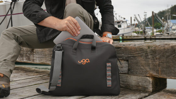 A man sitting on a wooden dock beam slides a silver laptop into a black ugo Messenger waterproof bag with orange uZIP drysuit-style zipper, with fishing boats in the marina behind him.