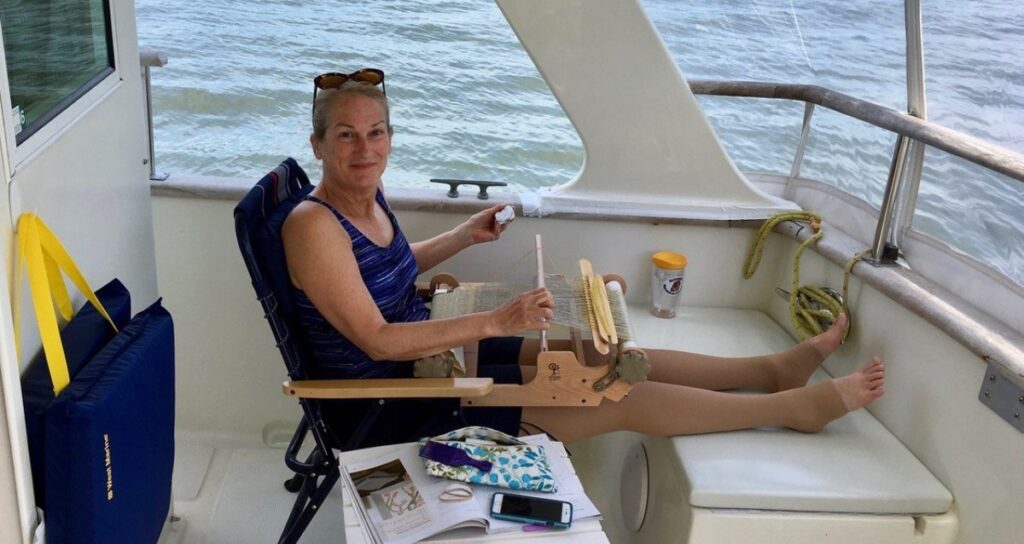 Woman cruising with lymphedema aboard a trawler, legs elevated with compression garment, weaving on a portable loom