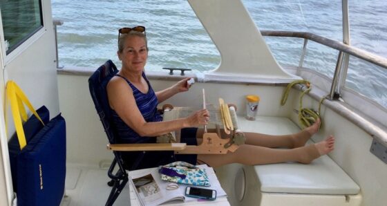 Woman cruising with lymphedema aboard a trawler, legs elevated with compression garment, weaving on a portable loom