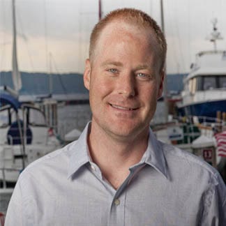 Man with red hair and grey shirt standing in front of boats